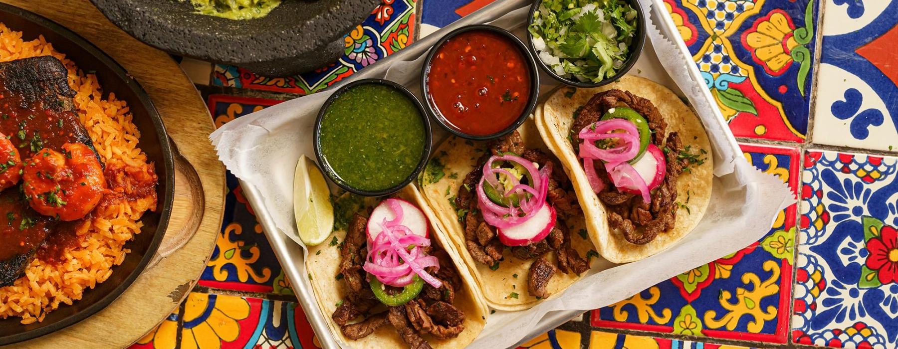 A tray of three tacos with salsa, guacamole, and rice on a colorful table.